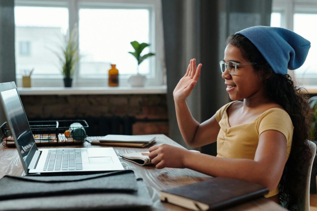 A young girl waves at her laptop during a video call in a cozy home office setting.